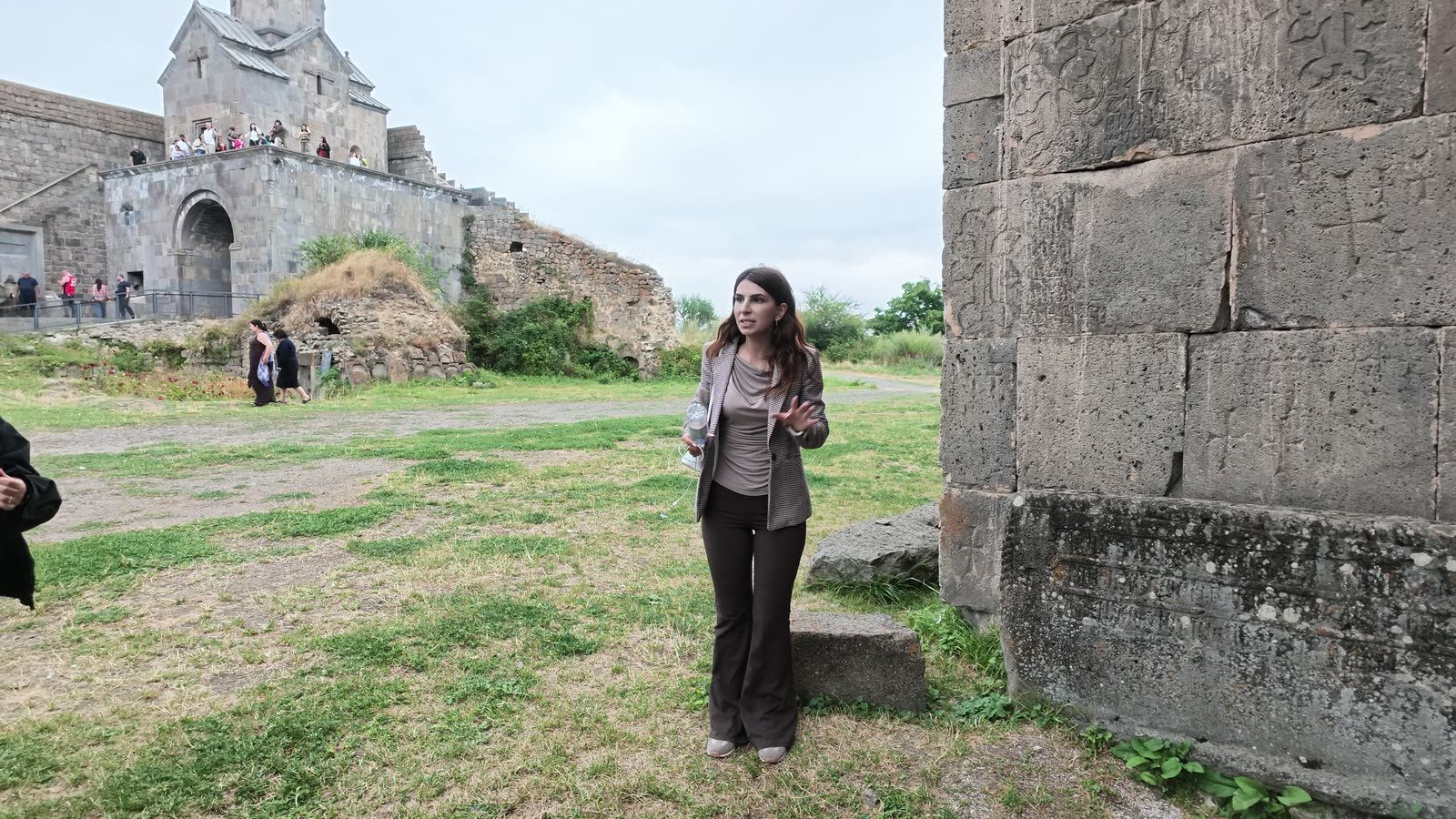 Karina, a guide, speaking beside the carved wall at Tatev monastery, the church behind her.